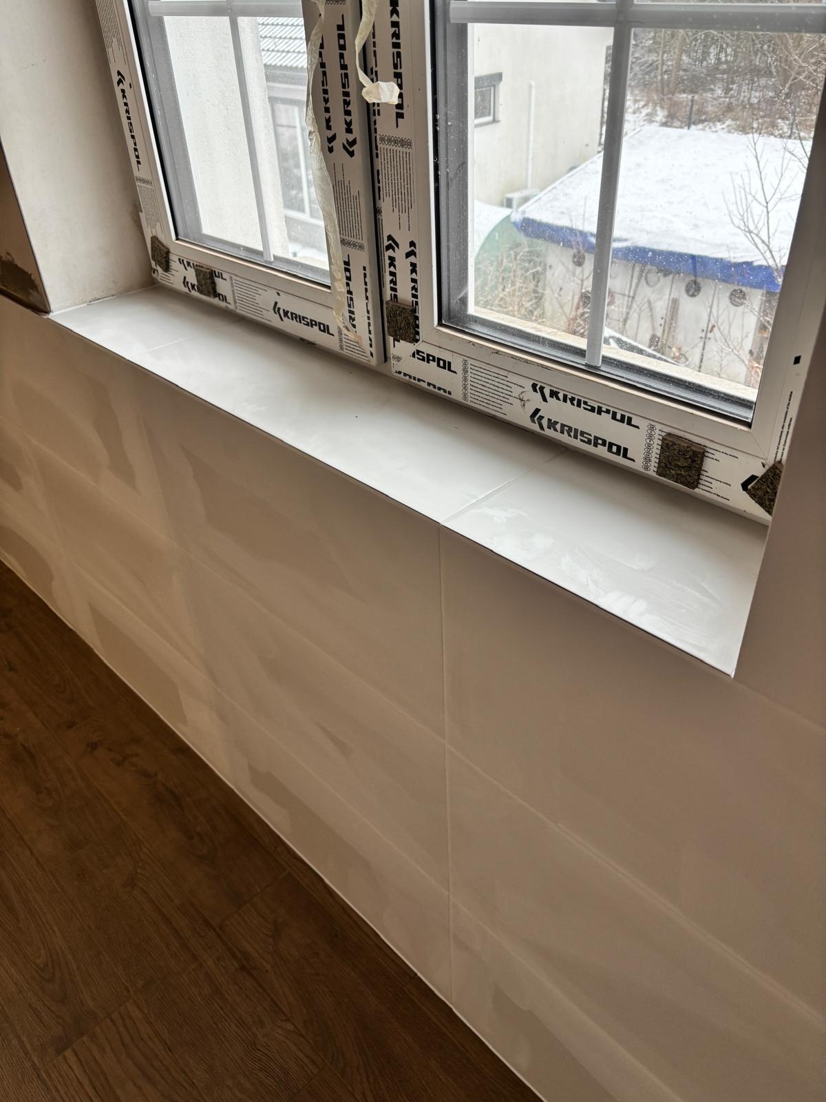 Błyszczące, kremowe płytki ścienne ułożone pod oknem z białymi ramami. Widoczny fragment drewnianej podłogi. Zewnętrzny widok na ośnieżony krajobraz.