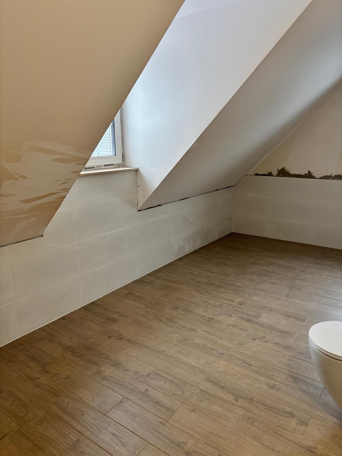 Remont łazienki na poddaszu: ułożone płytki imitujące drewno na podłodze, białe płytki na ścianach do połowy wysokości, ślady po szpachlowaniu na skosach.