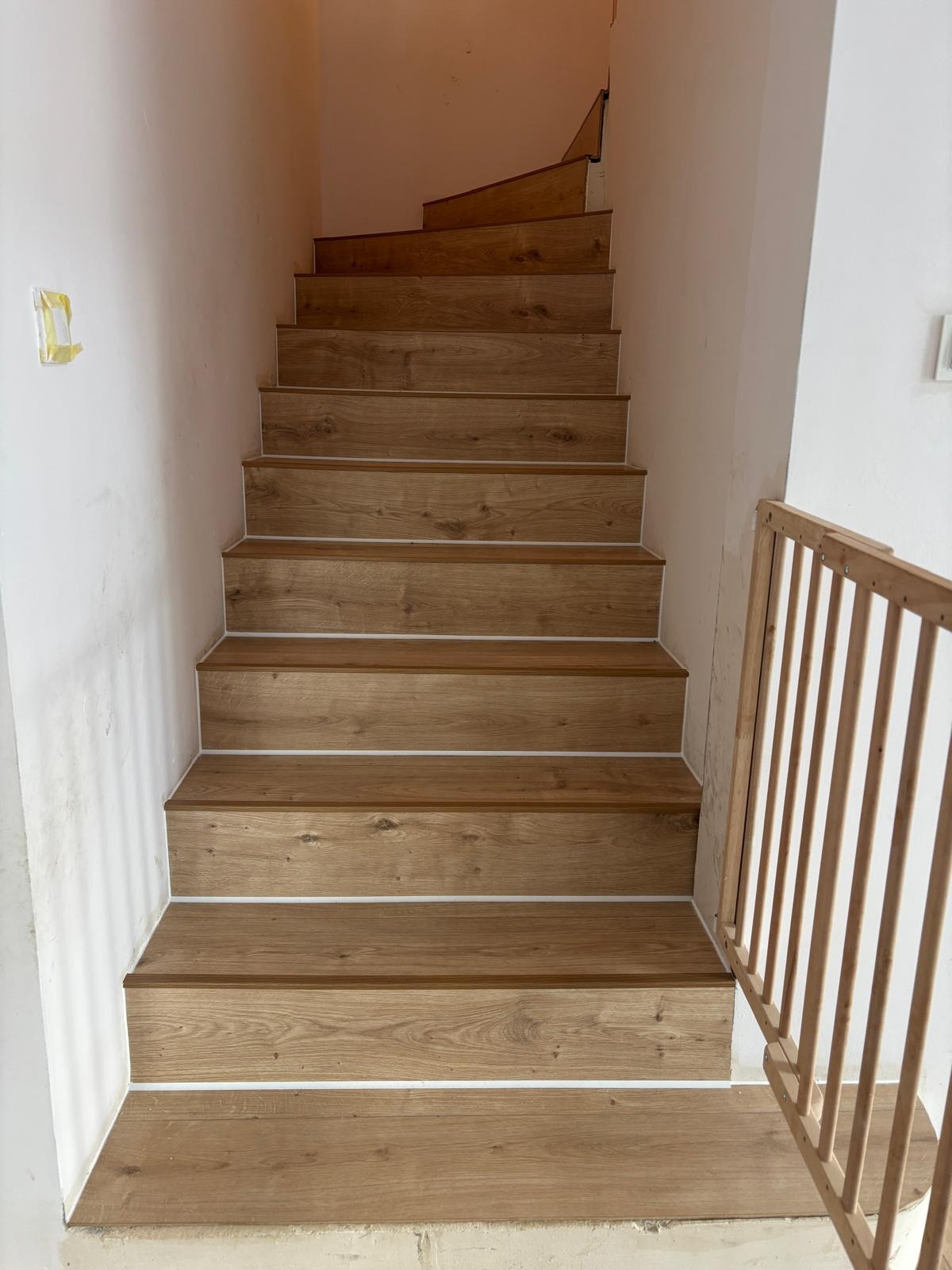 Schody wyłożone panelami drewnopodobnymi z białymi listwami przypodłogowymi, widok z dołu. Częściowo widoczna barierka zabezpieczająca.