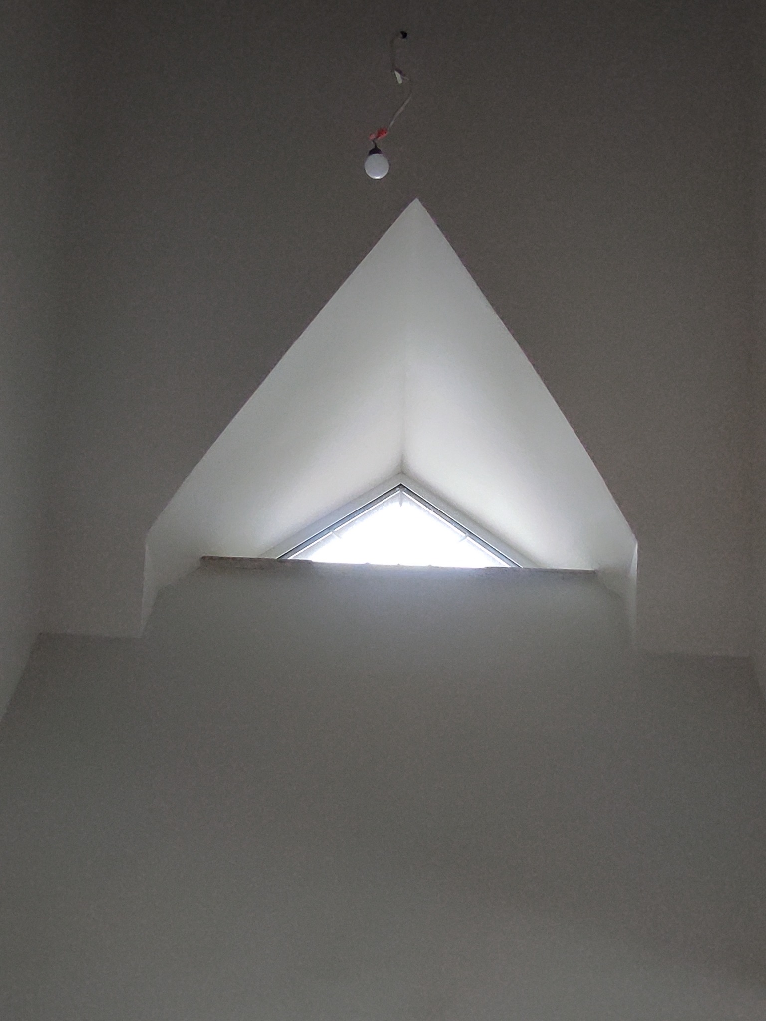 Biała ściana z trójkątnym oknem na poddaszu, widok z dołu. Widoczne gładzie i surowy stan wykończenia. U góry zwisa żarówka.