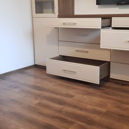 Rystol sp.z oo - Biało-brązowa szafa na wymiar z otwartymi szufladami i przeszkloną witryną, na tle drewnianej podłogi i telewizora. Widok z dołu, podkreślający przestronność wnętrza.