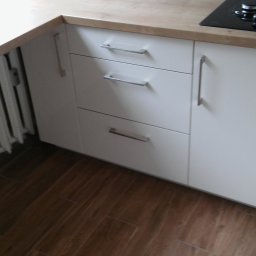 Rystol sp.z oo - Białe szafki kuchenne z drewnianym blatem i metalowymi uchwytami, widoczne brązowe płytki podłogowe i fragment grzejnika. Minimalistyczny design.