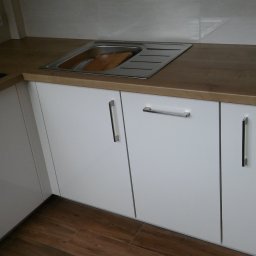 Rystol sp.z oo - Białe szafki kuchenne z drewnianym blatem i metalowym zlewozmywakiem, widok z bliska na fragment zabudowy kuchennej z płytkami w tle.