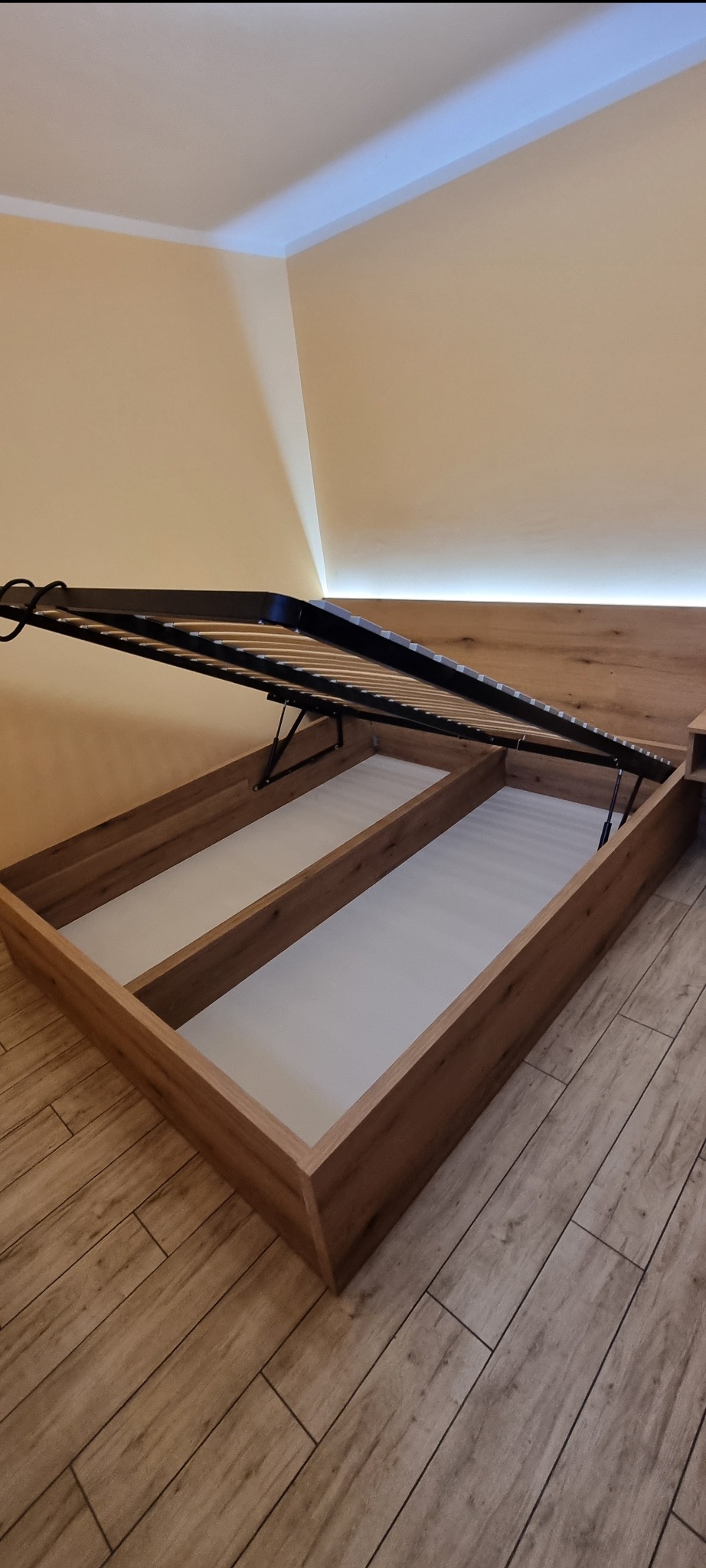 Otwarta rama łóżka z mechanizmem podnoszenia, ukazująca przestrzeń do przechowywania. Widoczne drewniane wykończenie i białe dno skrzyni. Podłoga z paneli imitujących drewno.