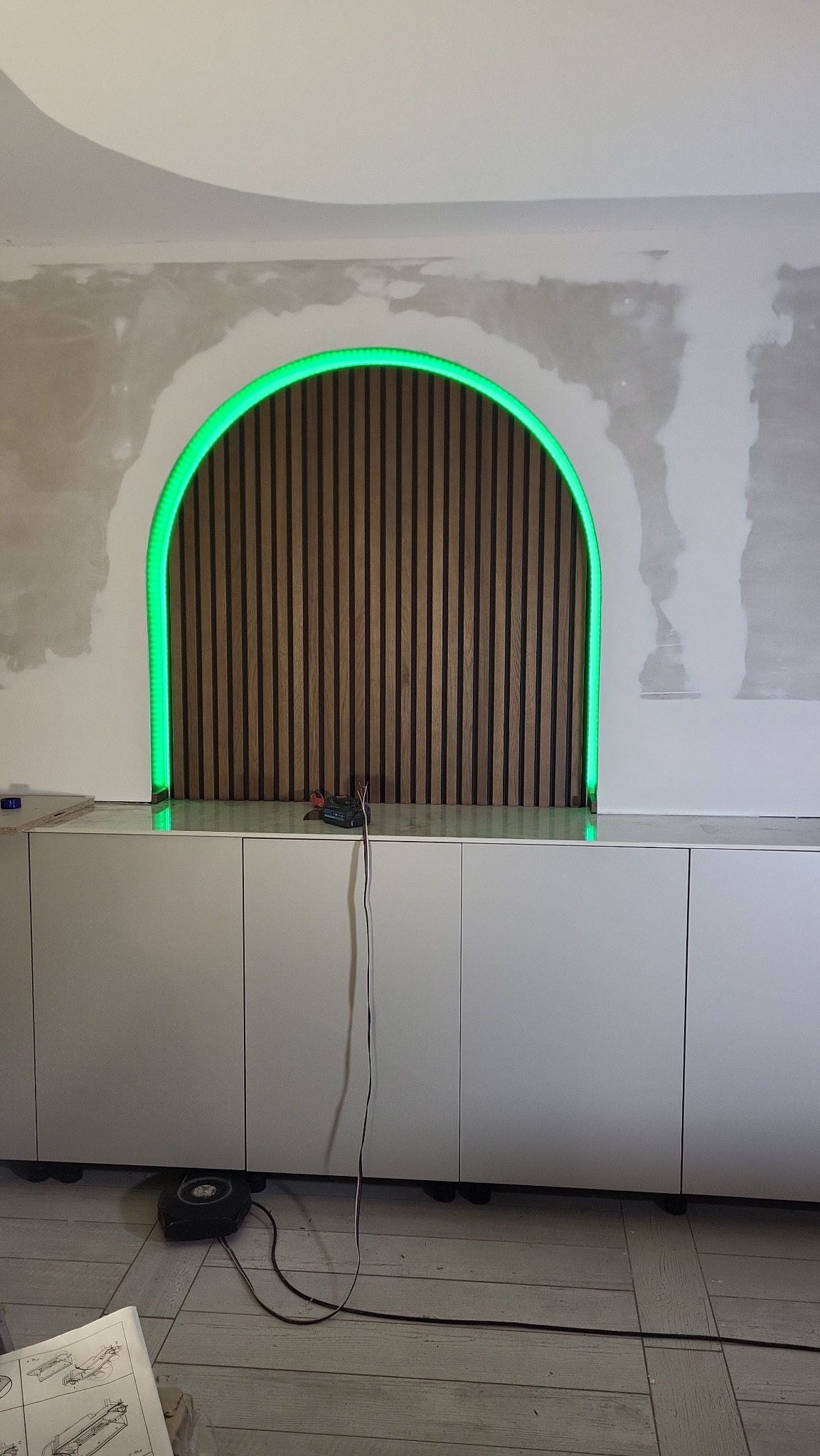 Narożna szafka z drewnianą ścianką działową w kształcie łuku, podświetloną zielonym neonem. Ściana w trakcie wykończenia, widoczne ślady po szpachlowaniu.