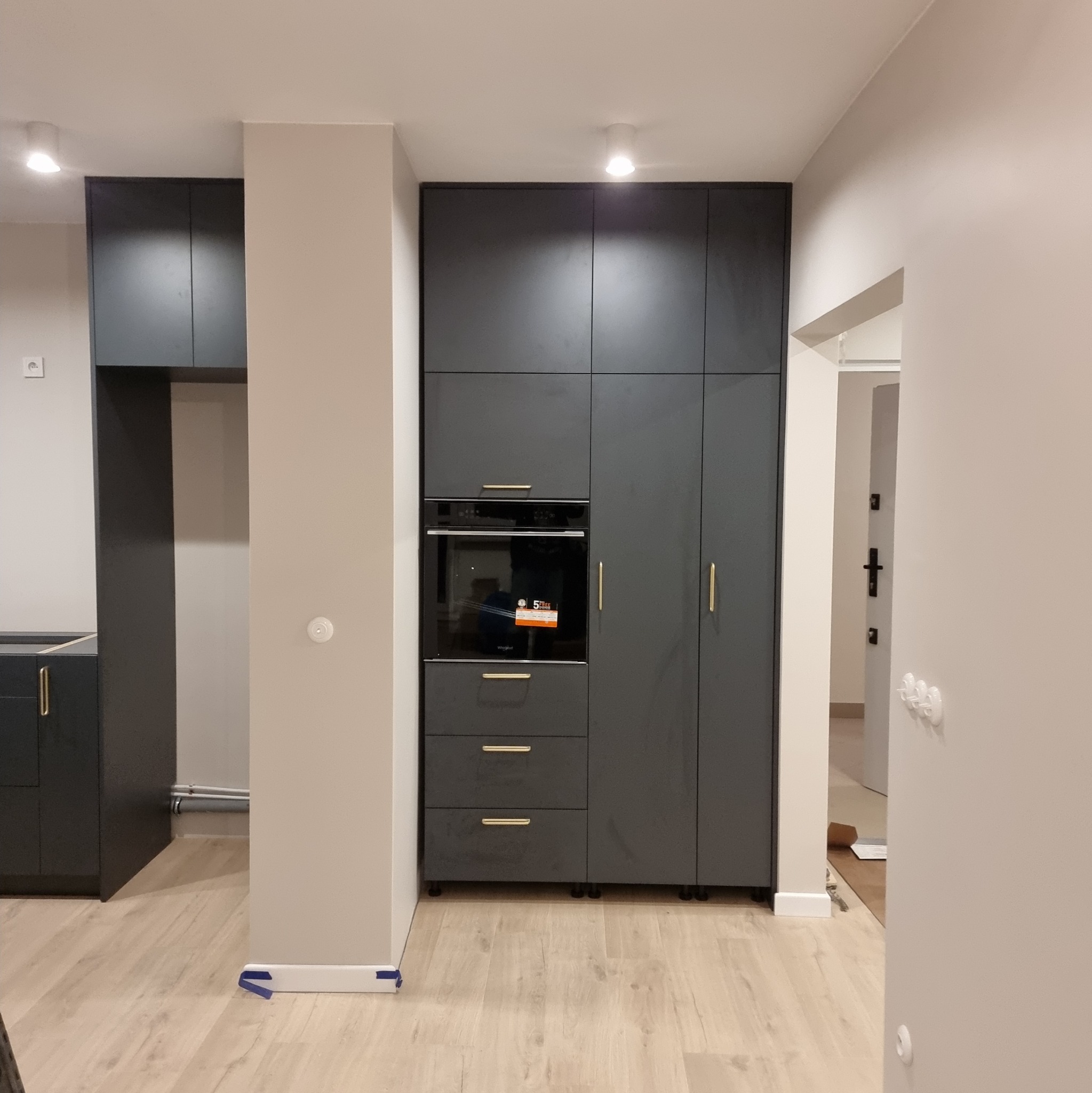 Ciemnoszara zabudowa kuchenna z piekarnikiem i złotymi uchwytami, na jasnej podłodze, widok z perspektywy wejścia do pomieszczenia. Minimalistyczny design.