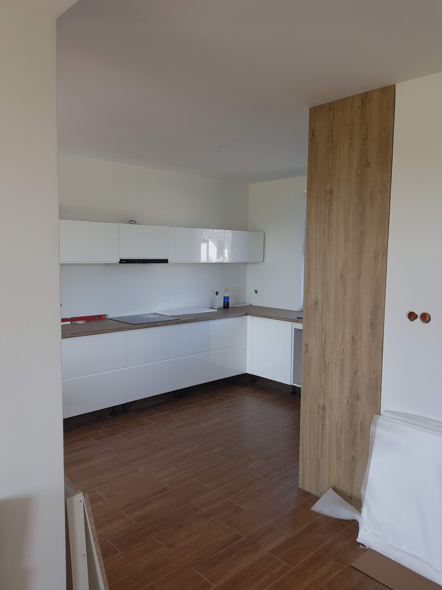 Nowoczesna, biała kuchnia z drewnianym panelem ściennym i brązową podłogą. Widoczne elementy montażowe i poziomica, sugerujące etap wykończenia.