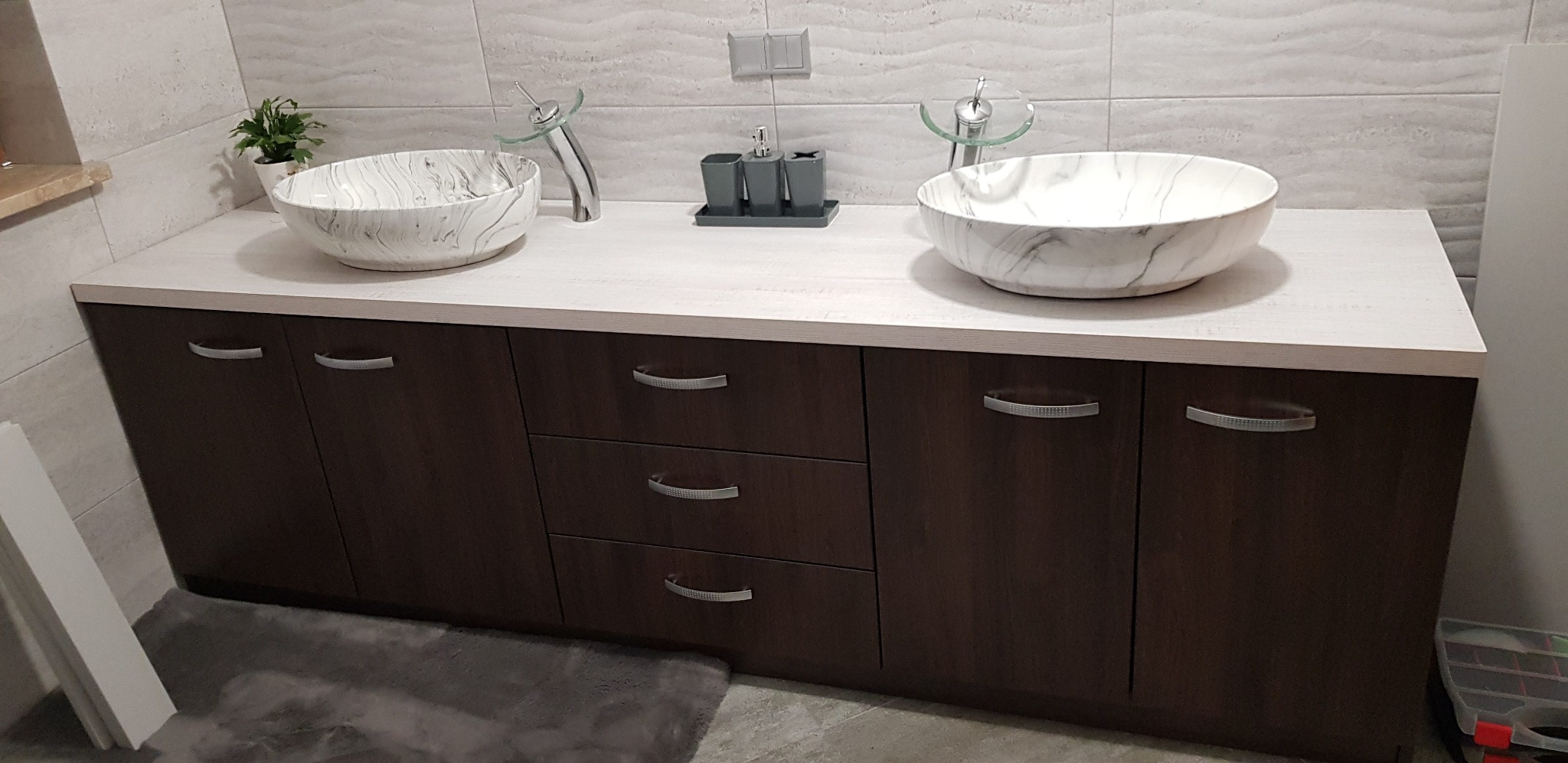 Nowoczesna łazienka z dwoma umywalkami w marmurowym wzorze, osadzonymi na szafce z ciemnego drewna. Minimalistyczne baterie i akcesoria dopełniają aranżację.