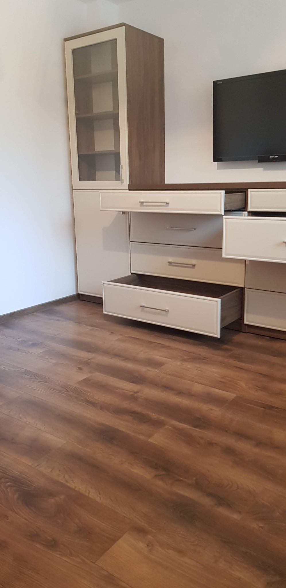 Biało-brązowa szafa na wymiar z otwartymi szufladami i przeszkloną witryną, na tle drewnianej podłogi i telewizora. Widok z dołu, podkreślający przestronność wnętrza.