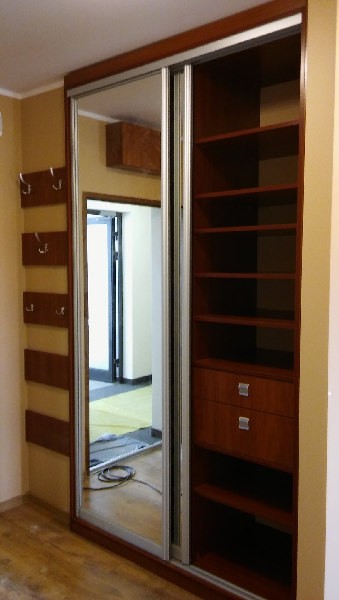 Brązowa szafa wnękowa z lustrzanymi drzwiami przesuwnymi, z widocznymi półkami i szufladami, zamontowana w pokoju z drewnianą podłogą i wieszakami na ścianie.