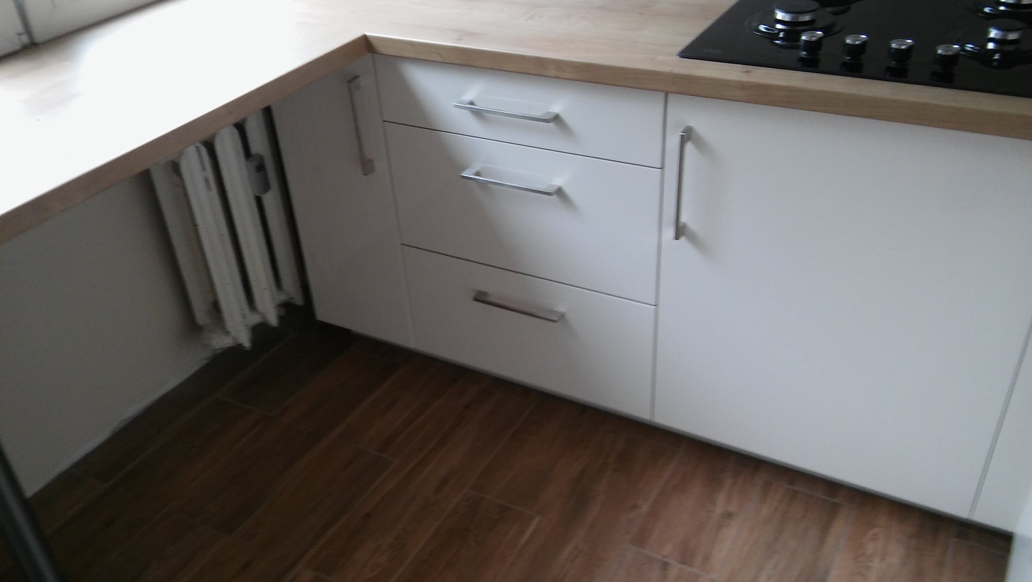 Białe szafki kuchenne z drewnianym blatem i metalowymi uchwytami, widoczne brązowe płytki podłogowe i fragment grzejnika. Minimalistyczny design.