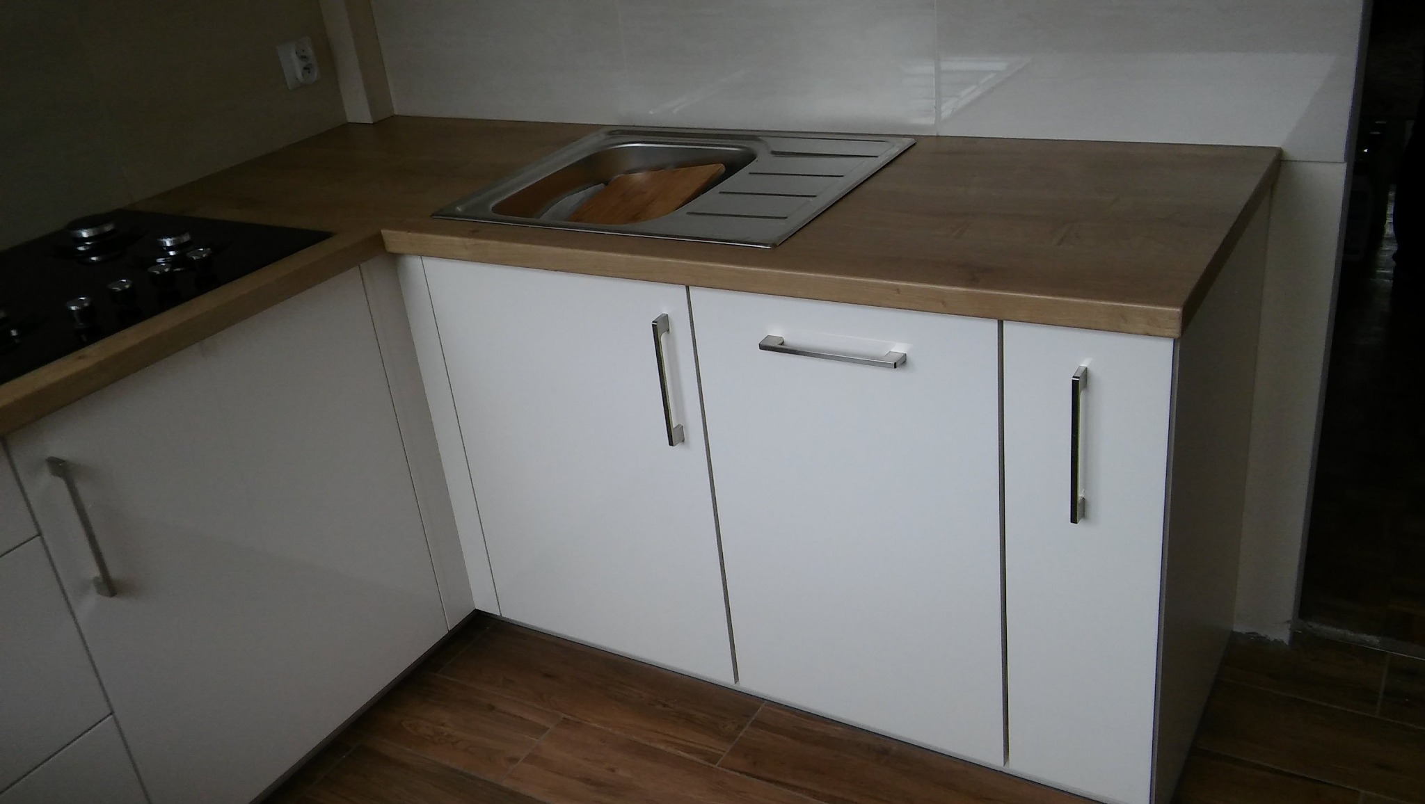 Białe szafki kuchenne z drewnianym blatem i metalowym zlewozmywakiem, widok z bliska na fragment zabudowy kuchennej z płytkami w tle.