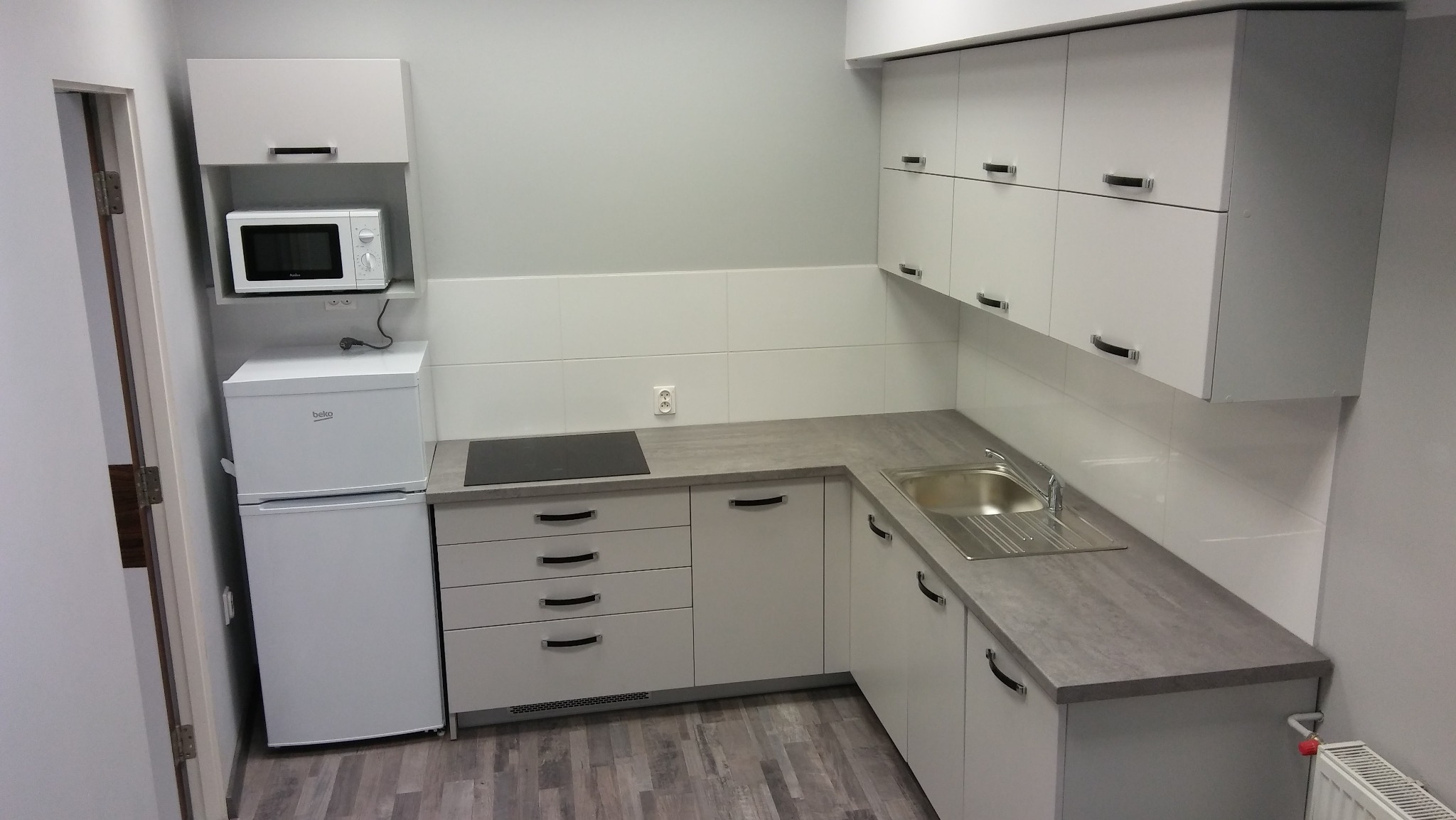 Jasna zabudowa kuchenna z szarym blatem i sprzętem AGD w Toruniu. Minimalistyczny design, widok na szafki górne i dolne, zlew oraz płytę indukcyjną.