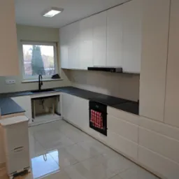 Nowoczesna kuchnia z białymi szafkami, czarnym blatem i armaturą, widok na okno z drzewem, płytki podłogowe imitujące kamień, czerwona ściereczka w piekarniku.