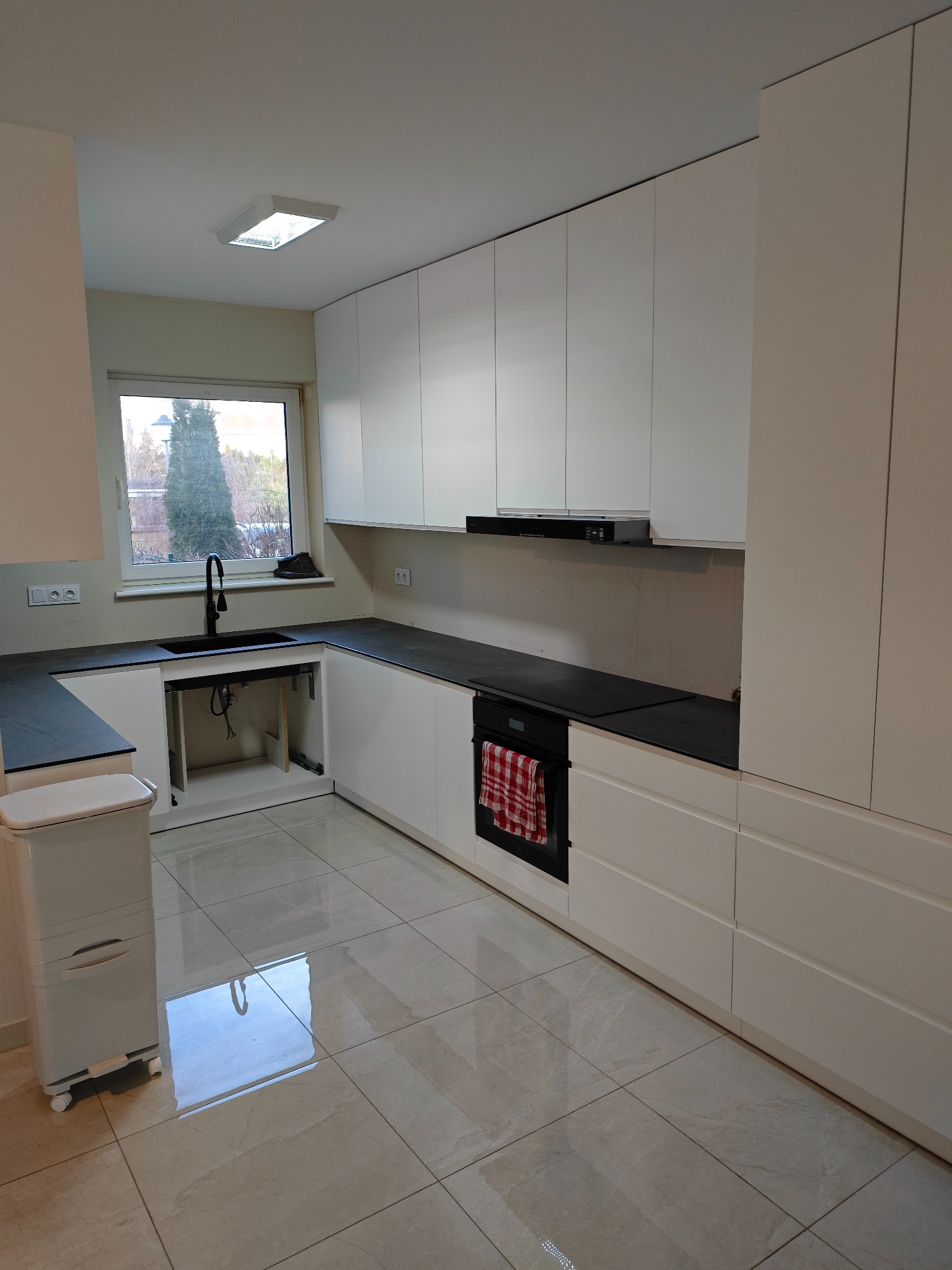 Nowoczesna kuchnia z białymi szafkami, czarnym blatem i armaturą, widok na okno z drzewem, płytki podłogowe imitujące kamień, czerwona ściereczka w piekarniku.