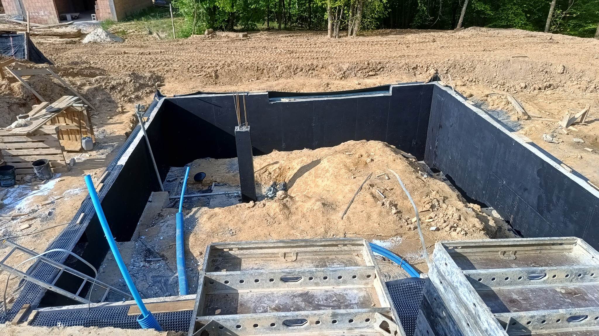 Wykonana izolacja pionowa fundamentów z widocznymi rurami drenarskimi i szalunkami, zasypana częściowo piaskiem.