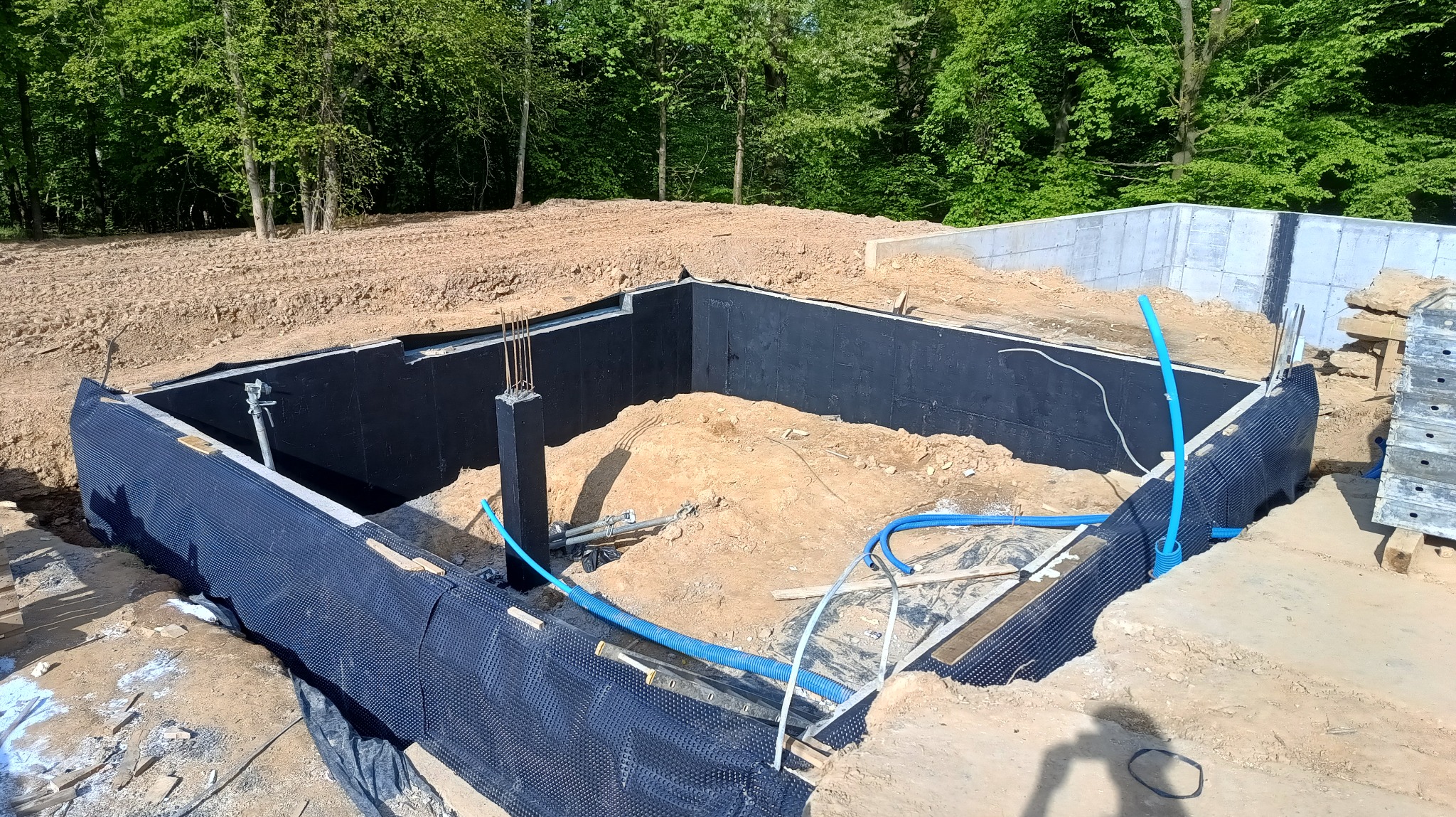 Wykop pod fundamenty z izolacją przeciwwilgociową, zbrojenie pionowe w narożniku, niebieskie rury instalacyjne wewnątrz wykopu.