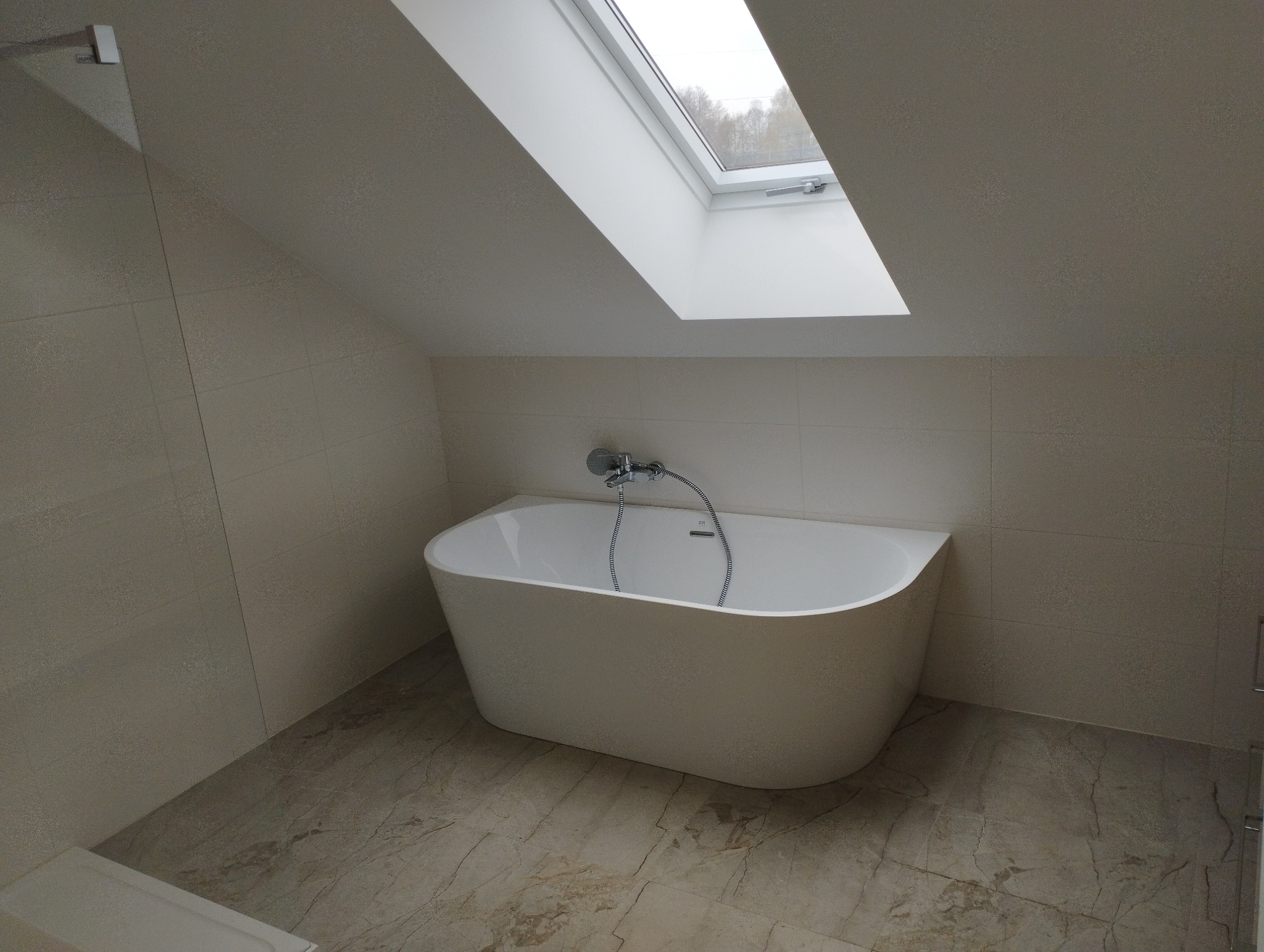 Nowoczesna, wolnostojąca wanna w łazience na poddaszu z oknem dachowym, wykończonej beżowymi płytkami imitującymi kamień.