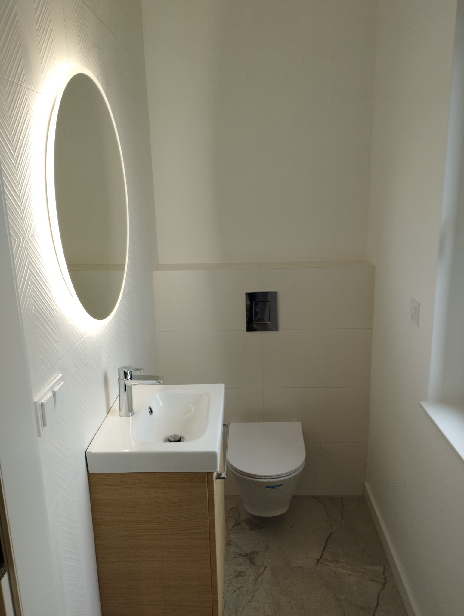 Nowoczesna łazienka w Dopiewie z podświetlanym, okrągłym lustrem nad umywalką z drewnianą szafką i podwieszaną toaletą, wykończona białymi płytkami z delikatnym wzorem i marmurową podłogą.