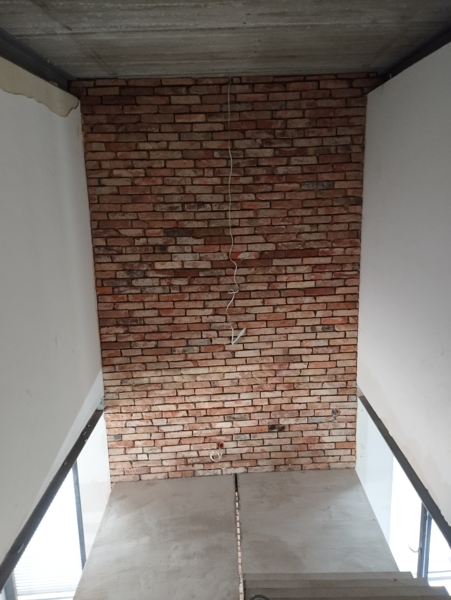 Ściana wyłożona cegłą klinkierową w trakcie prac wykończeniowych, widoczne przewody elektryczne i surowy betonowy strop