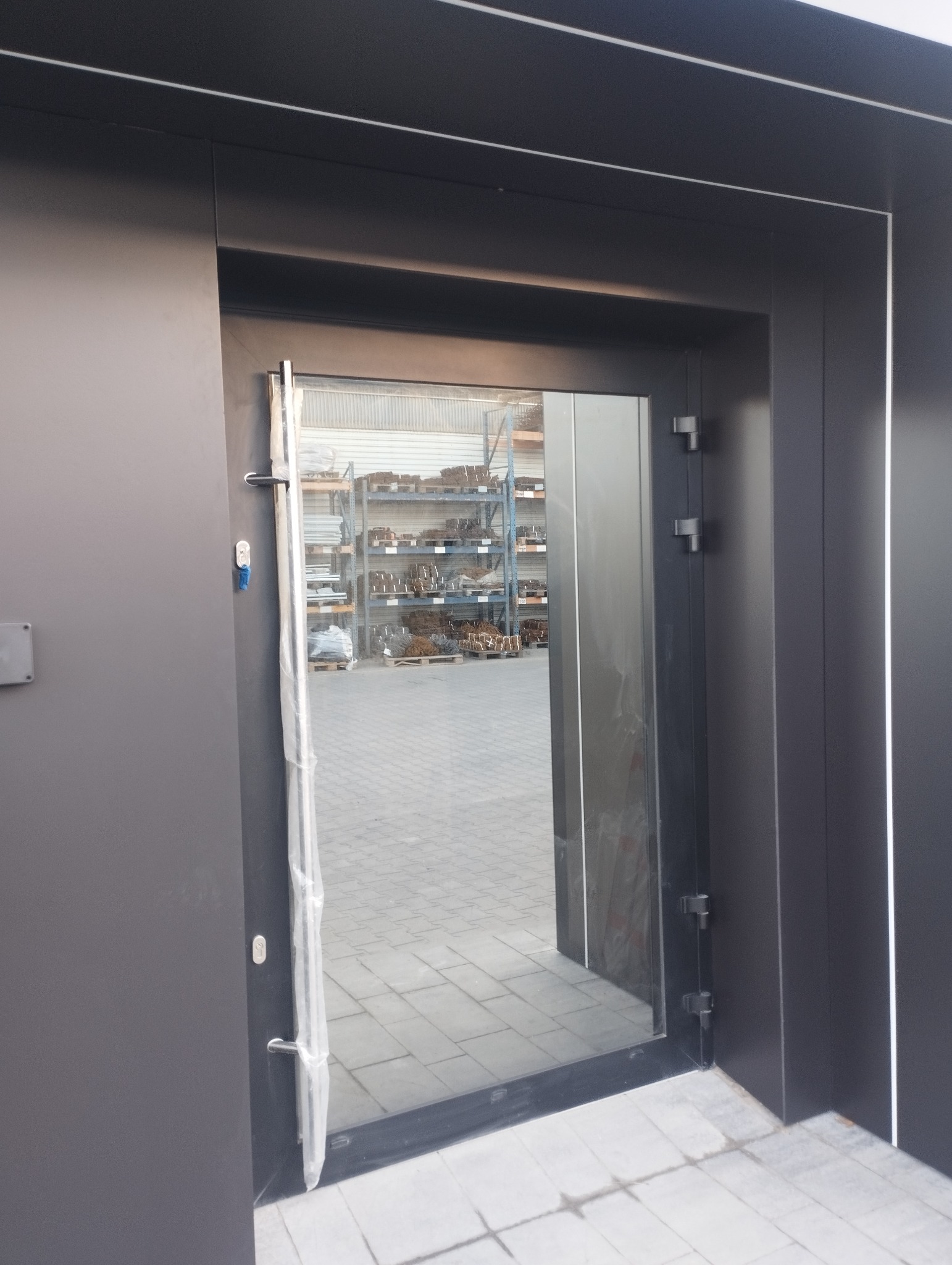 Ciemna, nowoczesna, przeszklona brama wejściowa z widoczną folią ochronną na ramie, przez którą widać magazyn z regałami i materiałami budowlanymi.