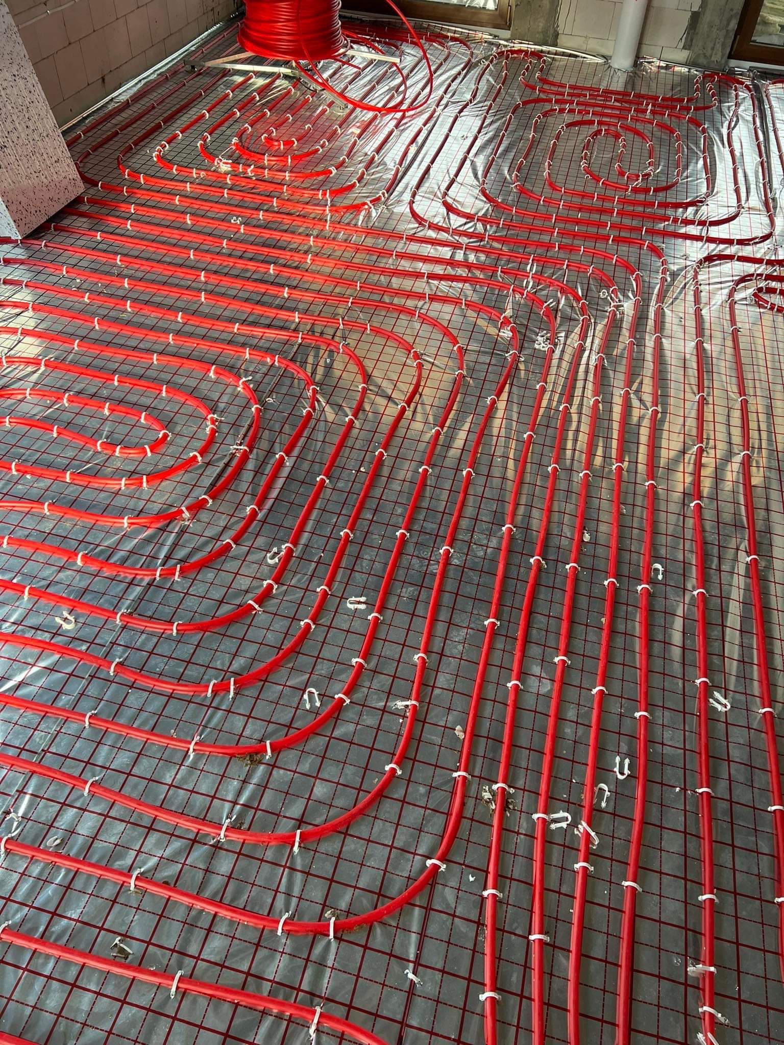 Czerwone rury ogrzewania podłogowego ułożone na siatce, mocowane białymi klipsami, widok z góry na etapie instalacji w surowym wnętrzu.