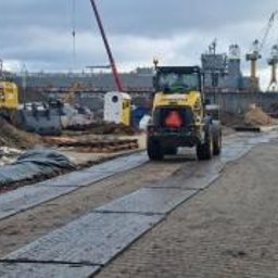 PRZEDSIĘBIORSTWO WIELOBRANŻOWE WIKTOR BORZYSZKOWSKI - Żółta ładowarka kołowa na placu budowy, poruszająca się po tymczasowej drodze z płyt z tworzywa sztucznego, w tle koparka i żurawie portowe pod pochmurnym niebem.