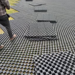 PRZEDSIĘBIORSTWO WIELOBRANŻOWE WIKTOR BORZYSZKOWSKI - Montaż systemu stabilizacji gruntu Terra-Grid E® 35 na trawiastym terenie; widoczne czarne, ażurowe płyty ułożone na powierzchni, z fragmentami trawy wystającymi przez otwory; w tle pracownicy...