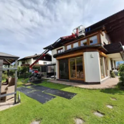 Prace na wysokości z użyciem podnośnika koszowego na trawniku zabezpieczonym czarnymi matami. Dwukondygnacyjny dom z drewnianymi elementami i licznymi oknami.