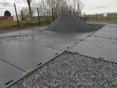 Czarne, plastikowe płyty tymczasowe ułożone na gruncie, tworzące drogę dojazdową, z widocznym stosem kruszywa w tle.