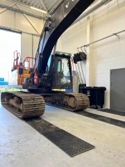 Ciężka koparka gąsienicowa wjeżdża na metalowe płyty ochronne wewnątrz hali warsztatowej, widoczne oświetlenie sufitowe i częściowo otwarte drzwi garażowe w tle.