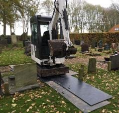 Mini koparka na cmentarzu, ustawiona na czarnej płycie drogowej, grzebiąca między nagrobkami.
