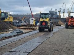 Żółta ładowarka kołowa na placu budowy, poruszająca się po tymczasowej drodze z płyt z tworzywa sztucznego, w tle koparka i żurawie portowe pod pochmurnym niebem.