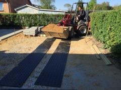 Miniładowarka z piaskiem na placu budowy, z czarnymi płytami ochronnymi na ziemi i kostką brukową w tle, przygotowanie terenu pod nawierzchnię.