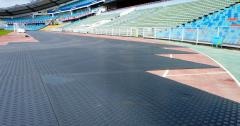 Nawierzchnia z czarnych płyt na bieżni lekkoatletycznej stadionu, widoczne trybuny w tle.