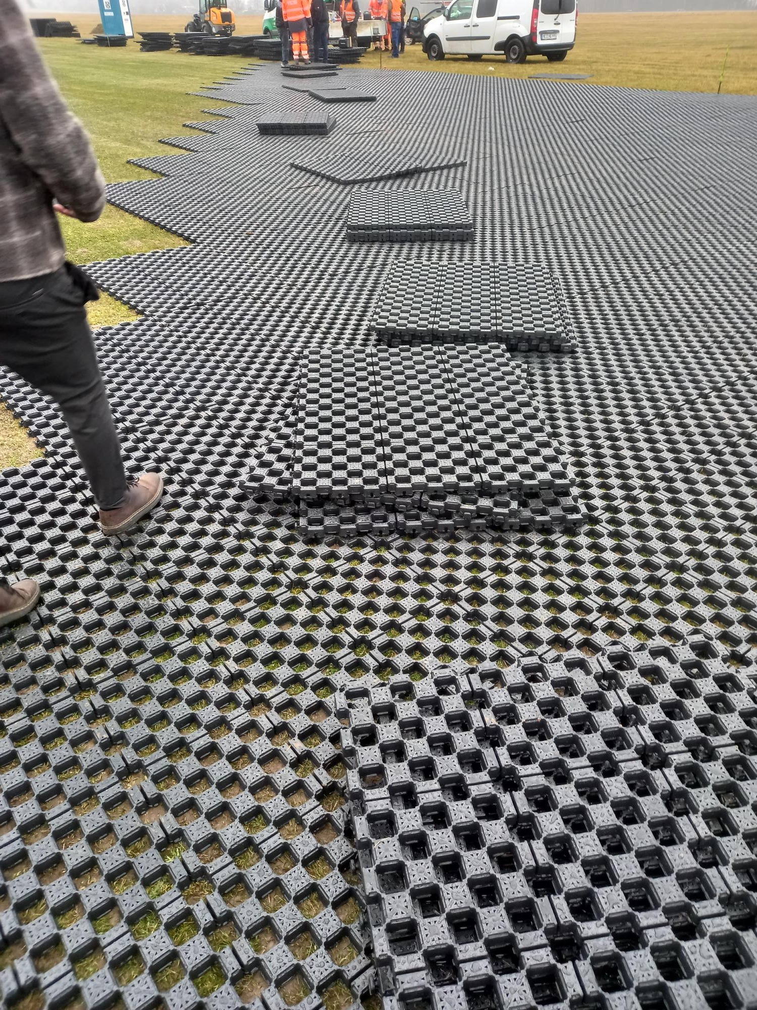 Montaż systemu stabilizacji gruntu Terra-Grid E® 35 na trawiastym terenie; widoczne czarne, ażurowe płyty ułożone na powierzchni, z fragmentami trawy wystającymi przez otwory; w tle pracownicy...