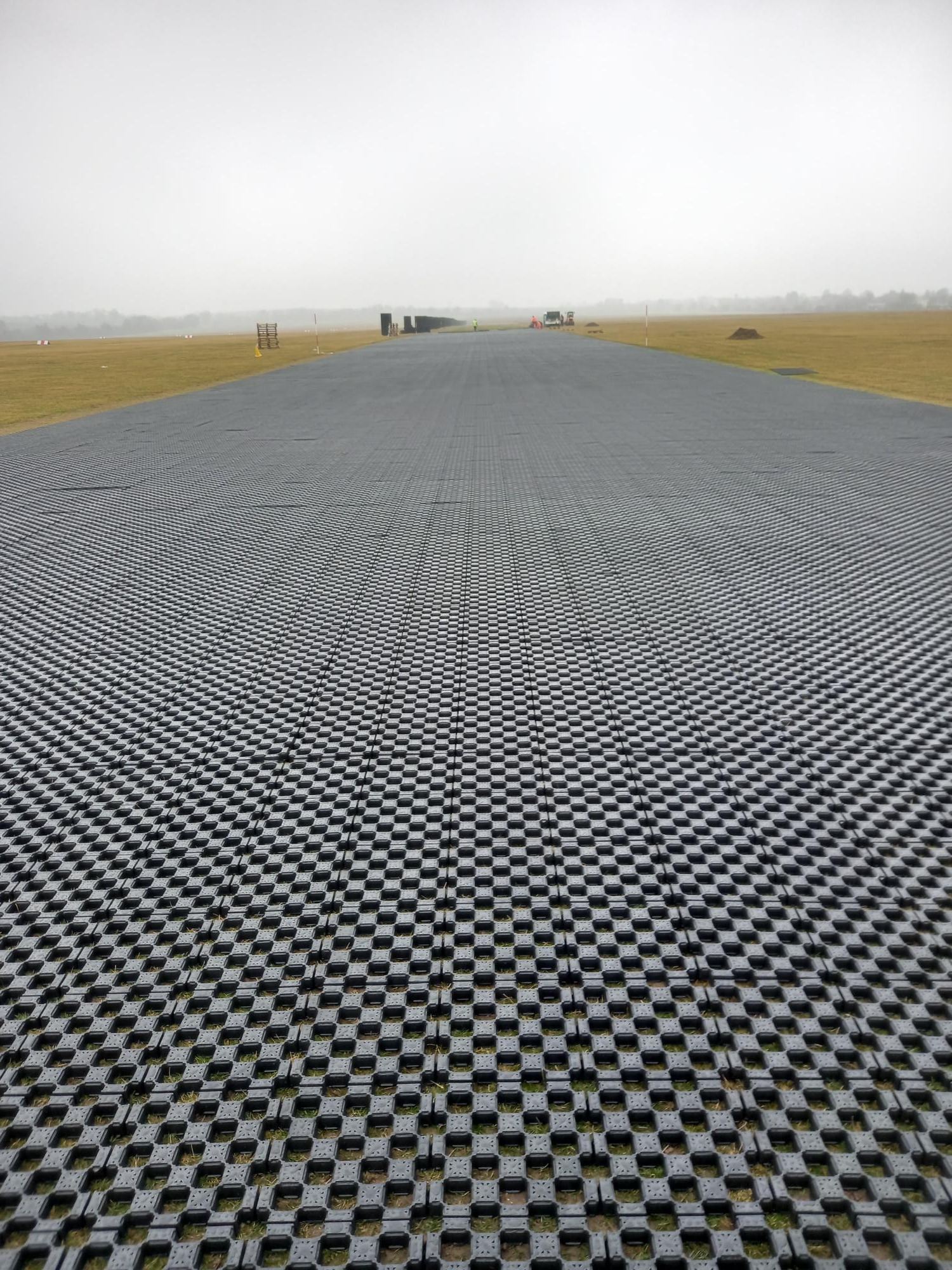 Rozległa powierzchnia wyłożona szarymi płytami ażurowymi Terra-Grid E® 35, widok z perspektywy ziemi, w tle słabo widoczne maszyny budowlane i drzewa, pochmurne niebo.
