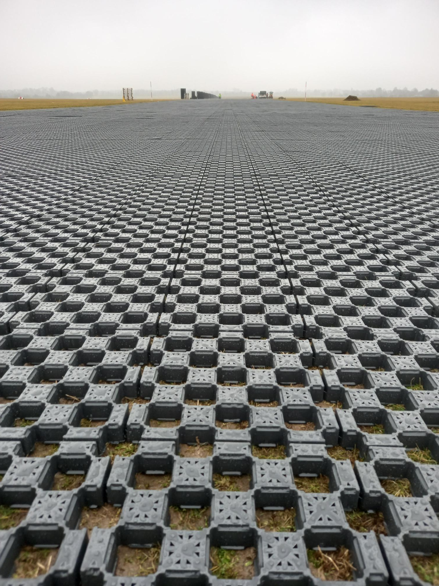 Rozległa powierzchnia pokryta szarymi, ażurowymi płytami drogowymi Terra-Grid E® 35, stabilizującymi grunt, z widoczną trawą i ziemią w otworach; w tle sprzęt budowlany i mglista przestrzeń.
