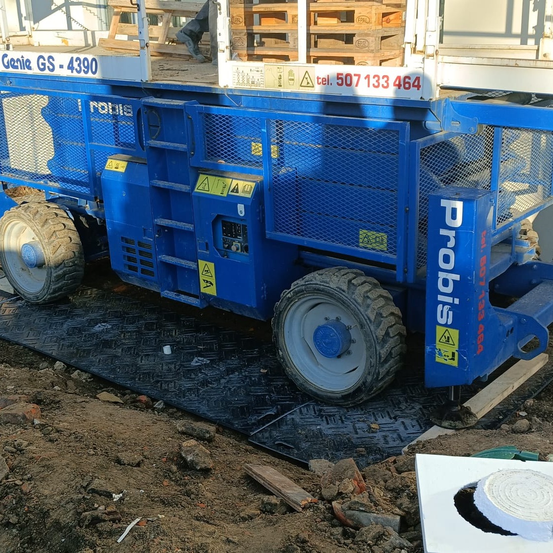 Niebieski podnośnik koszowy Genie GS-4390 stoi na czarnej, wzmocnionej nawierzchni. Widoczne palety w tle. Oznakowanie bezpieczeństwa na maszynie.