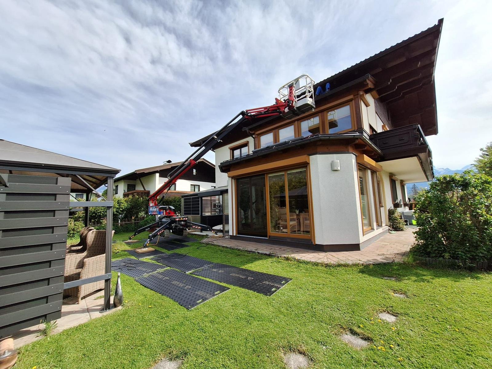 Prace na wysokości z użyciem podnośnika koszowego na trawniku zabezpieczonym czarnymi matami. Dwukondygnacyjny dom z drewnianymi elementami i licznymi oknami.