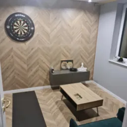 Pokój z wykończoną ścianą w jodełkę z drewnopodobnych paneli, tarczą do darta Winmau, szarą szafką RTV, stolikiem kawowym i zielonym fotelem.