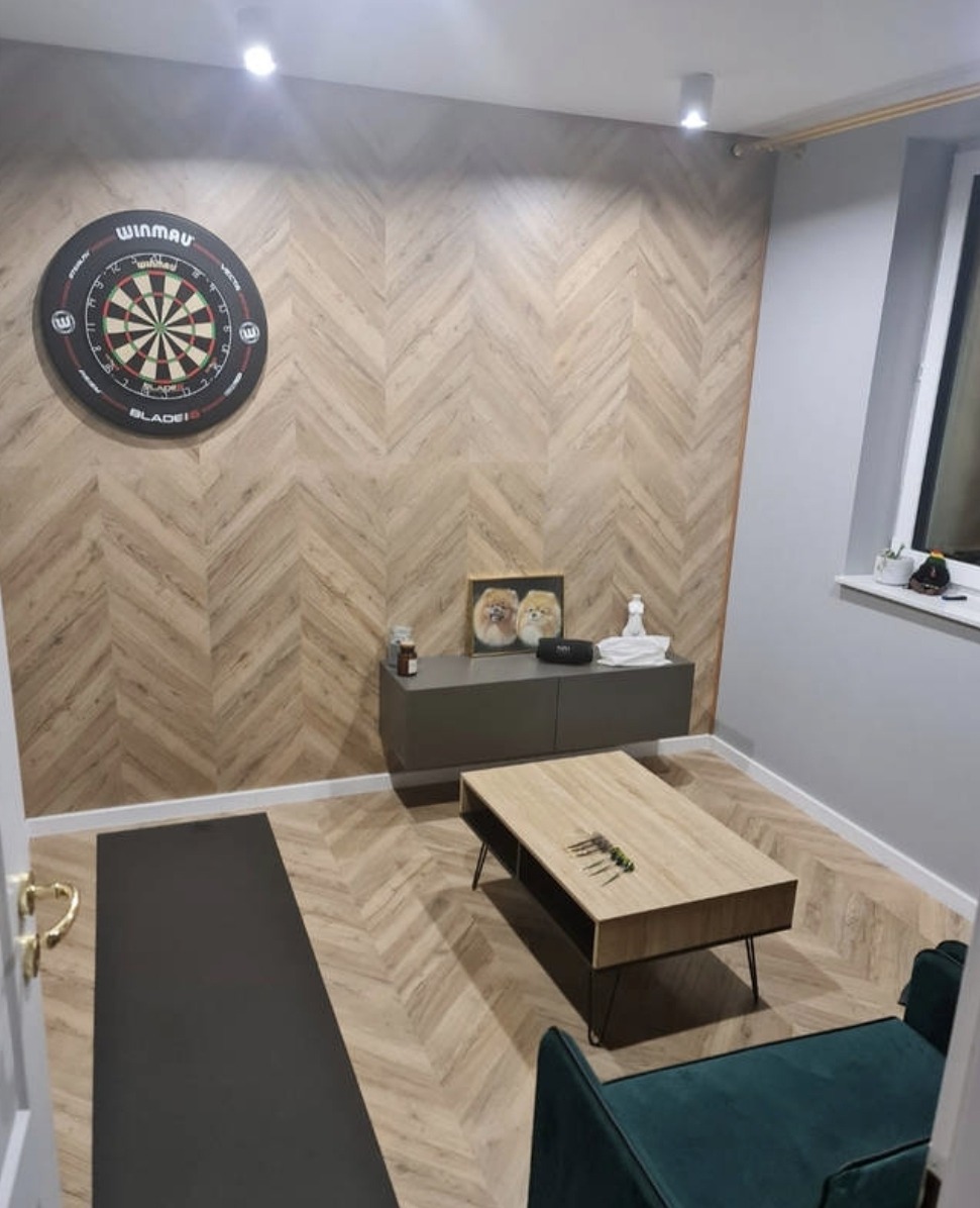 Pokój z wykończoną ścianą w jodełkę z drewnopodobnych paneli, tarczą do darta Winmau, szarą szafką RTV, stolikiem kawowym i zielonym fotelem.