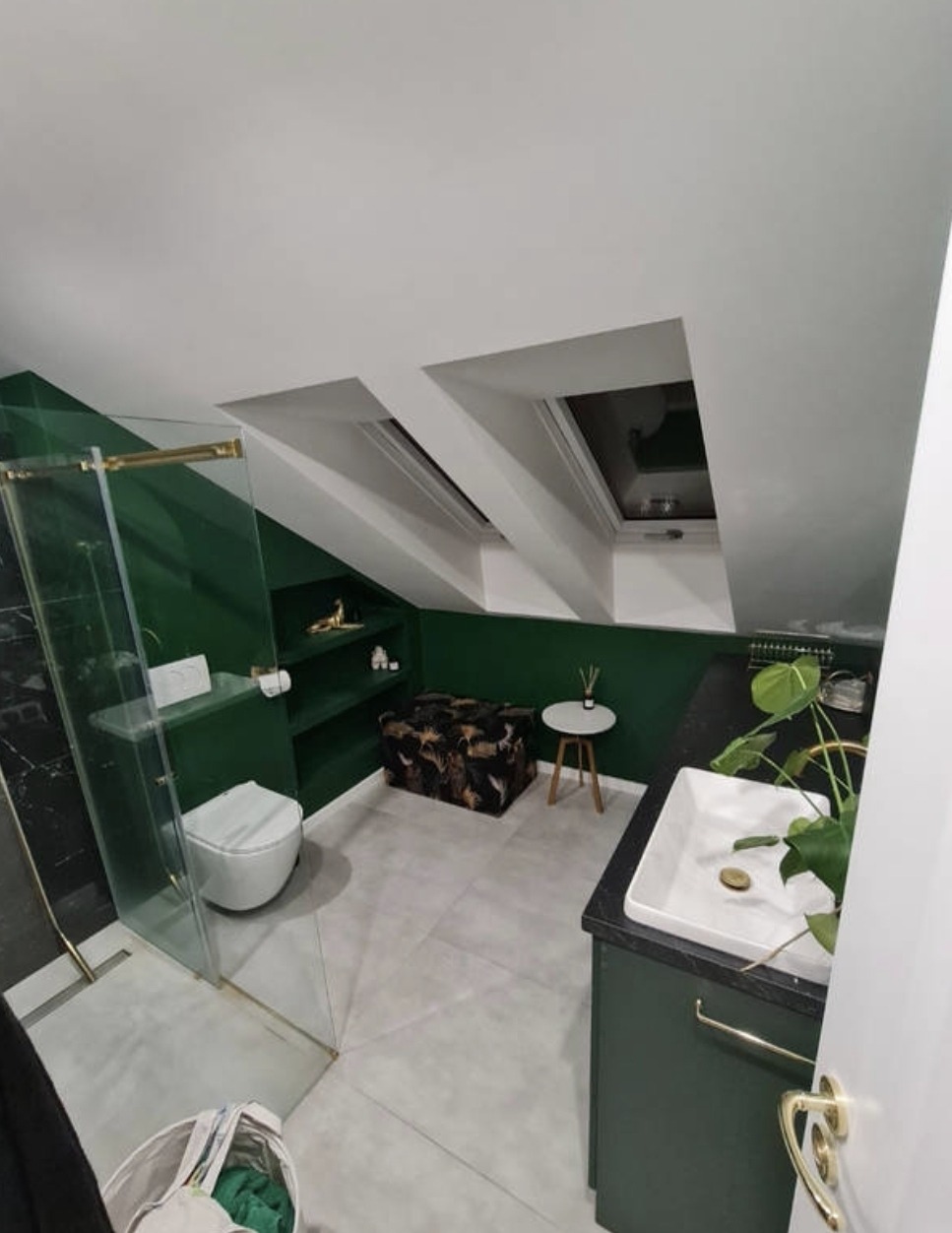 Nowoczesna łazienka na poddaszu ze skośnym sufitem, prysznicem ze złotymi detalami, zielonymi ścianami, szarą podłogą i oknami dachowymi.