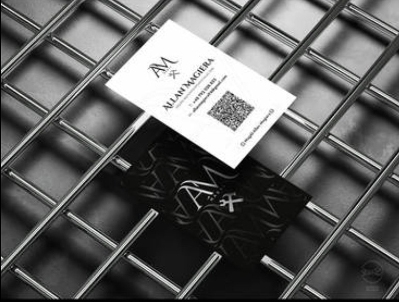 Dwie wizytówki firmy Allan Magiera leżące na metalowej kratce w ujęciu pod kątem, jedna biała z kodem QR, druga czarna z logo.