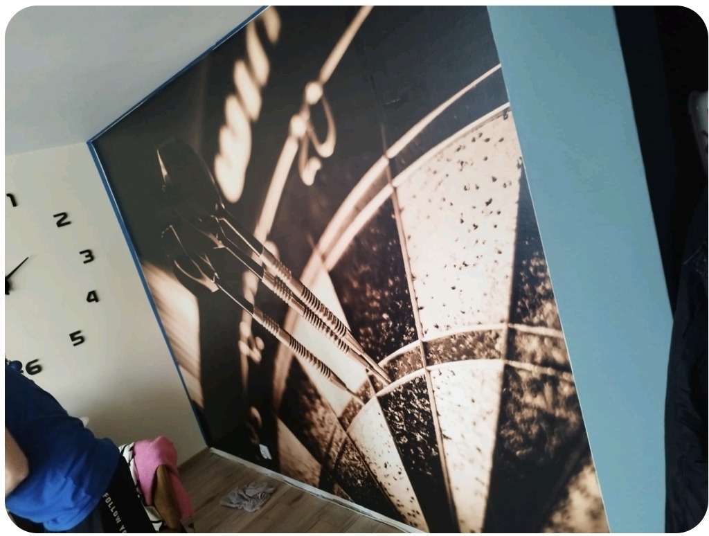 Fototapeta z motywem tarczy do darta i lotek w brązowych odcieniach, zamontowana na ścianie w pokoju, widoczny fragment ściany z zegarem.