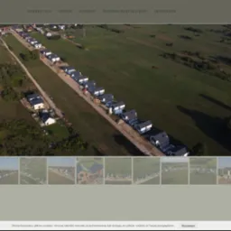 Widok z lotu ptaka na osiedle domów jednorodzinnych z ciemnymi dachami w trakcie budowy, otoczone zielonymi polami i lasami.