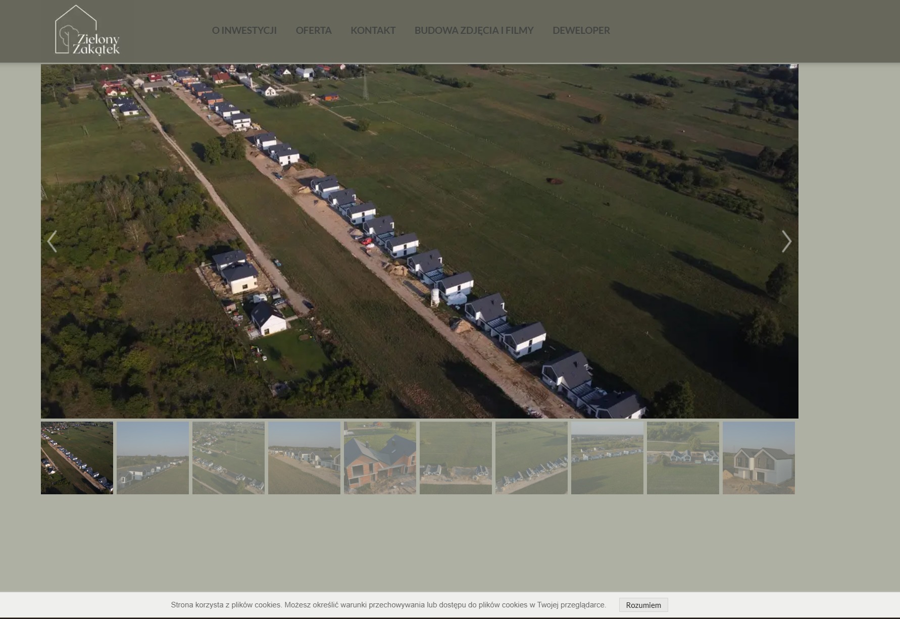 Widok z lotu ptaka na osiedle domów jednorodzinnych z ciemnymi dachami w trakcie budowy, otoczone zielonymi polami i lasami.