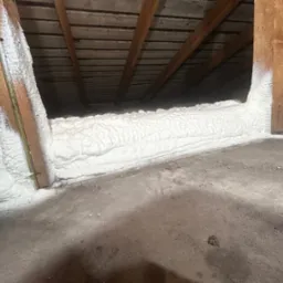 Poddasze ocieplone białą pianką PUR, widoczne drewniane krokwie i fragment betonowej podłogi z drobnymi zabrudzeniami.