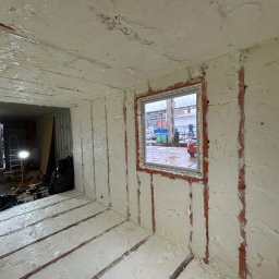 RAPIDFORCE SPÓŁKA Z OGRANICZONĄ ODPOWIEDZIALNOŚCIĄ - Wnętrze kontenera po natrysku izolacji pianką poliuretanową, widoczne nierównomierne pokrycie pianką na ścianach, suficie i podłodze, zarys konstrukcji kontenera widoczny spod pianki, okno...