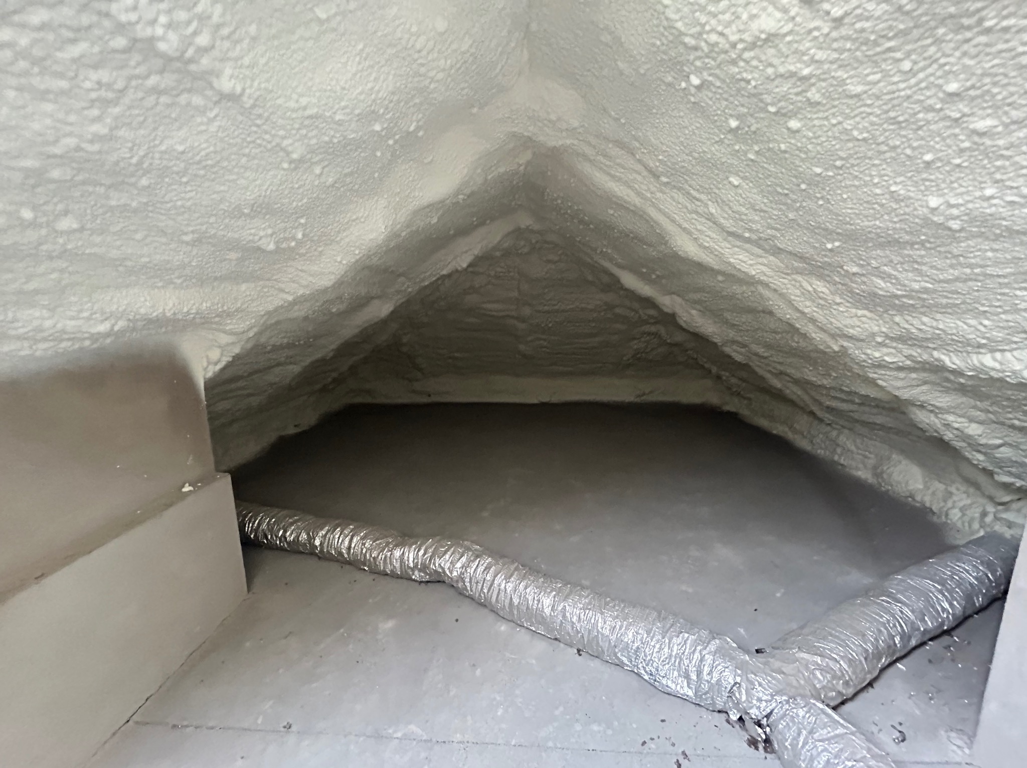 Poddasze z widoczną izolacją natryskową z pianki poliuretanowej i aluminiowym przewodem wentylacyjnym leżącym na podłodze.